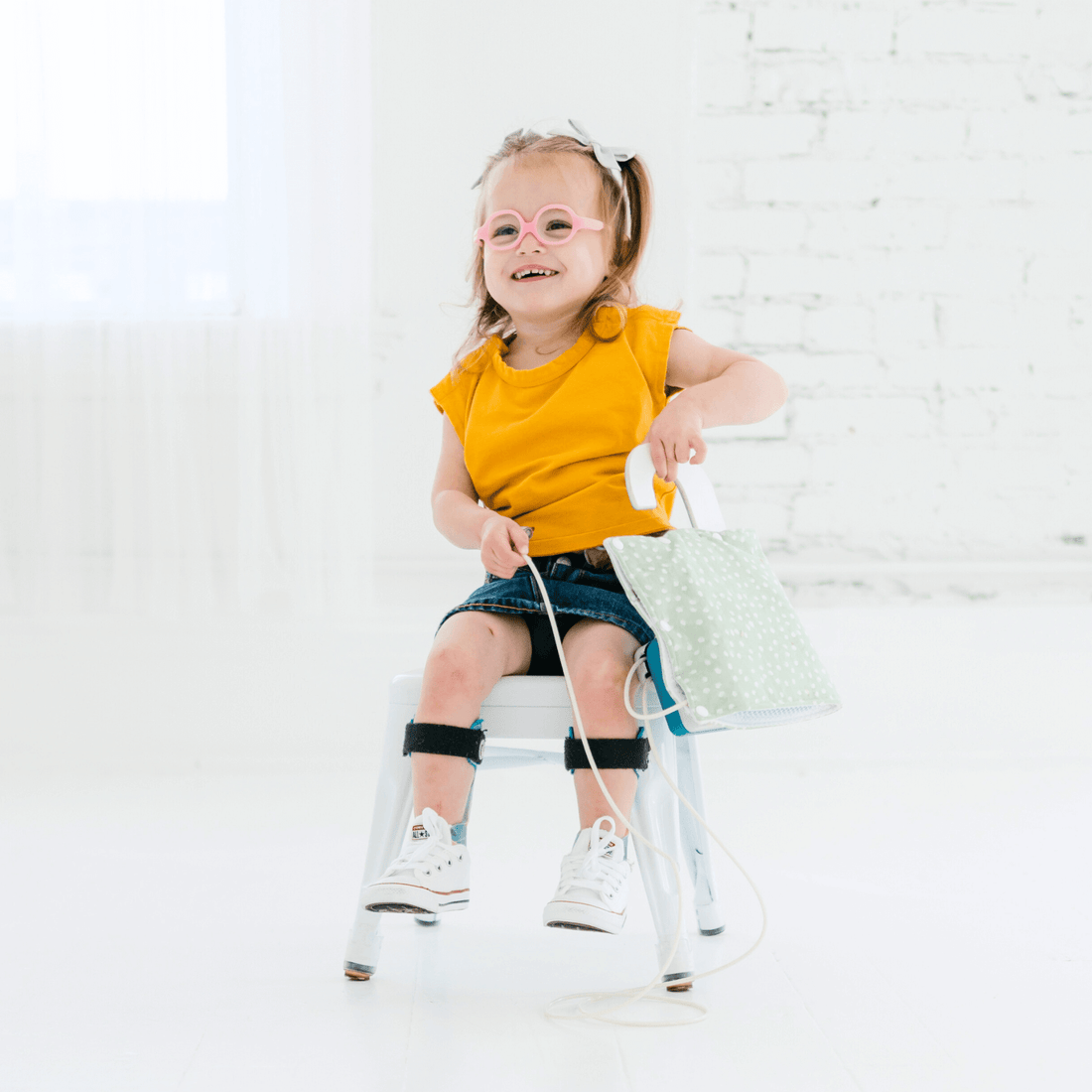 smiling-toddler-wearing-a-yellow-snappy-tee-and-holding-a-green-bag,-seated-on-a-chair-in-a-bright-room.
