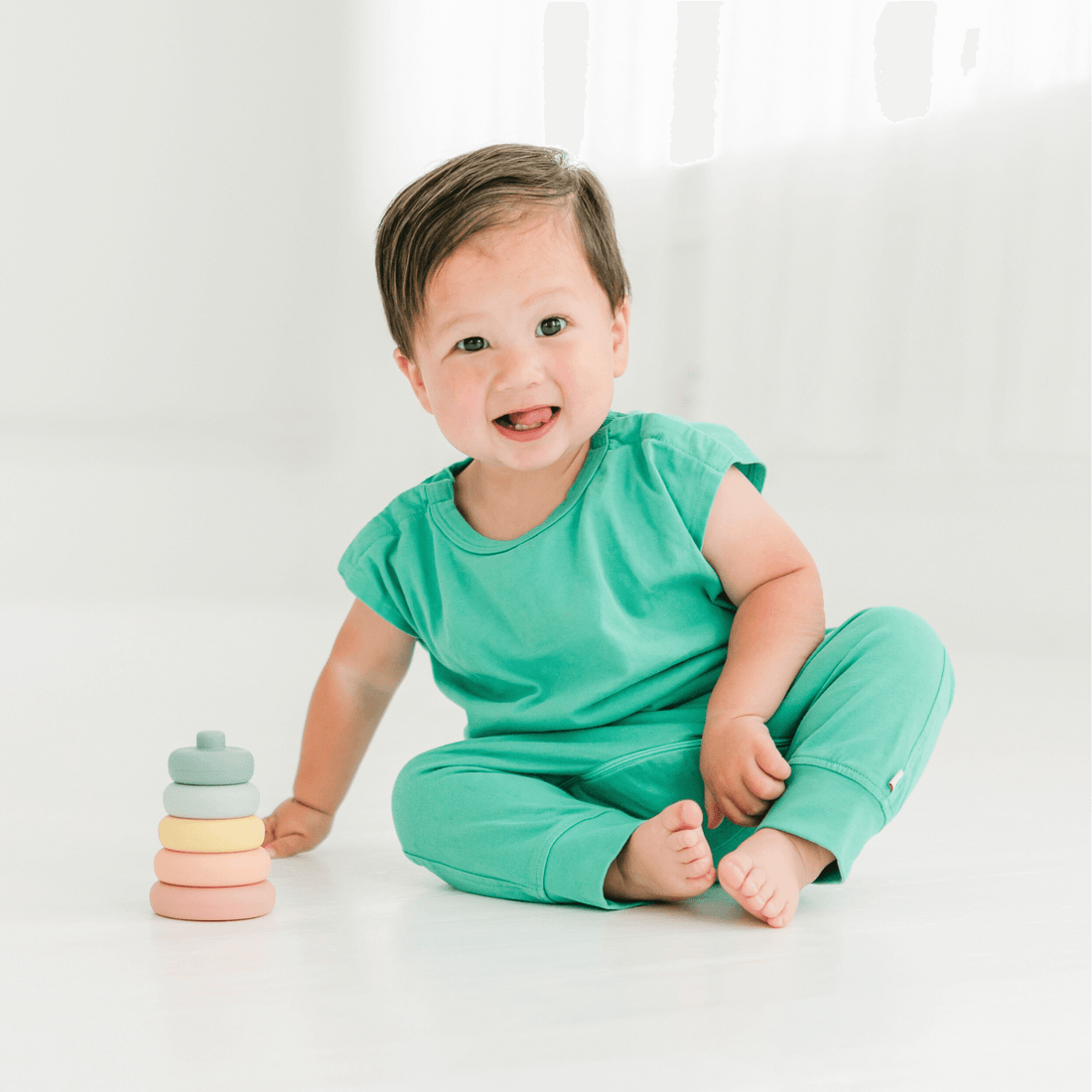 happy-toddler-in-mint-green-bodysuit-sitting-beside-colorful-stacking-toys,-showcasing-versatile-baby-clothing.